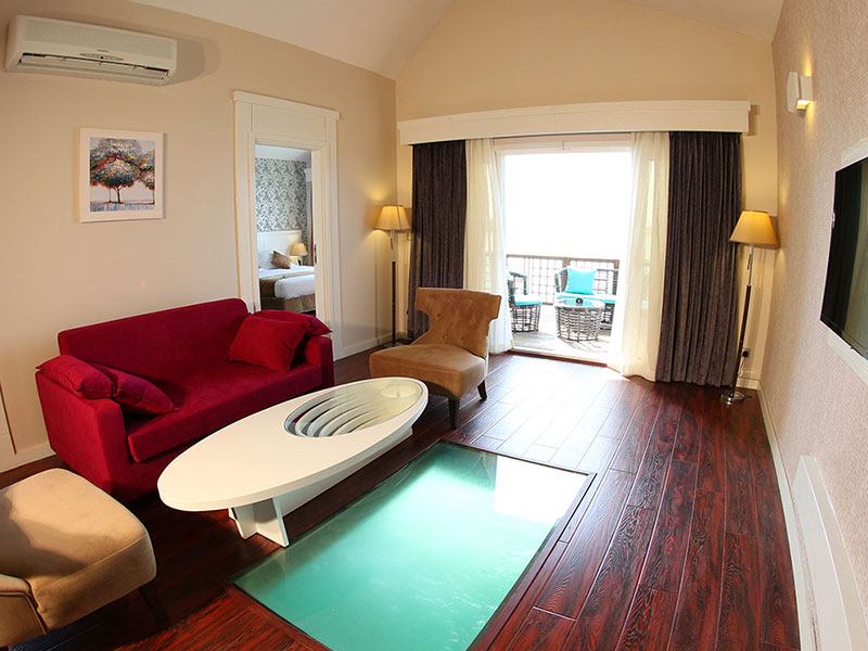 اتاق هتلی با کف شیشه ای به دریا و مبلمان قرمز و خردلی و میز سفید بیضی شکل