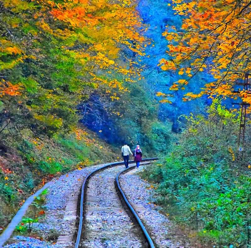 عبور دو رهگذر از روی ریل راه آهن سراسری ایران میان جاده های پاییزی با درختان زرد و سبز