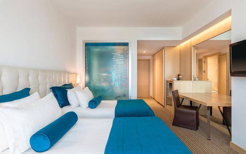 دو تخت دونفره با ملافه آبی و میز تحریر در اتاق هتل