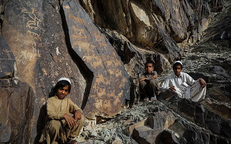 سه کودک با لباس محلی در میان صخره‌هایی با حکاکی های تاریخی