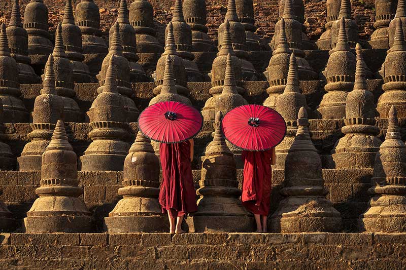 دو نفر با لباس و چتر قرمز روبروی معبدی در میانمار