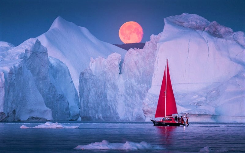 قایق بادبانی قرمز در کنار کوه‌های یخ گرینلند