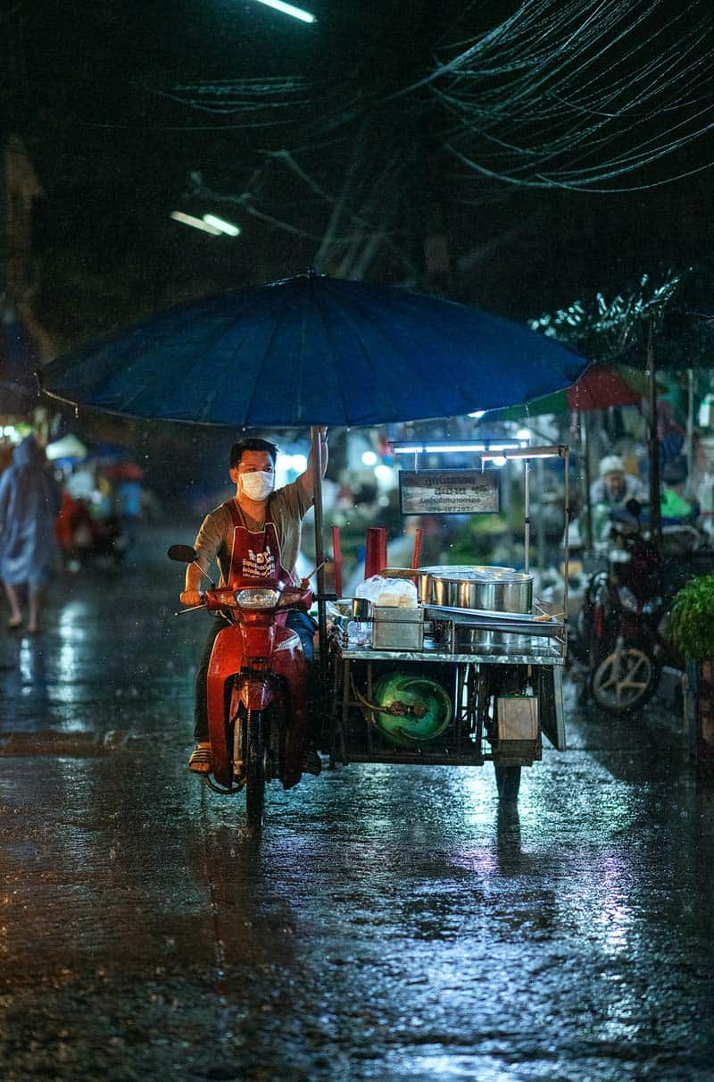 فروشنده مرد روی سه چرخه‌ای در خیابان بارانی شهر چیانگ مای