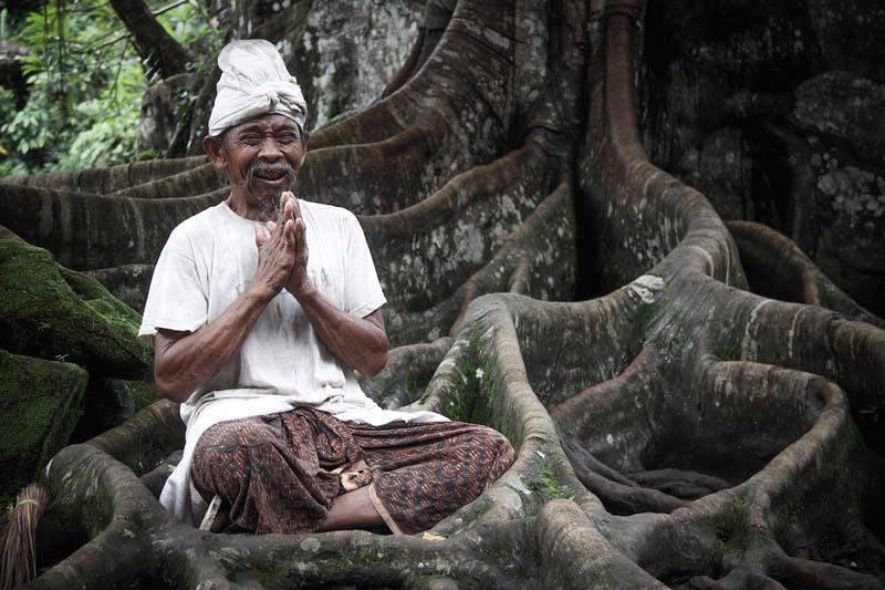 مرد آسیایی با لباس سفید در کنار درختی پر ریشه 