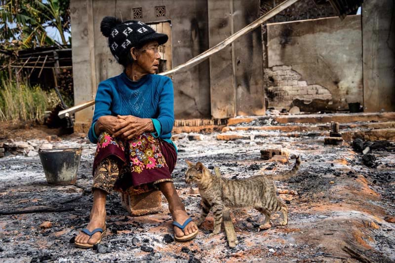 نشستن زنی در کنار یک گربه در مقابل خانه خود که از جنگ و درگیری بین ارتش میانمار و ارتش استقلال کاچین آسیب دیده است.