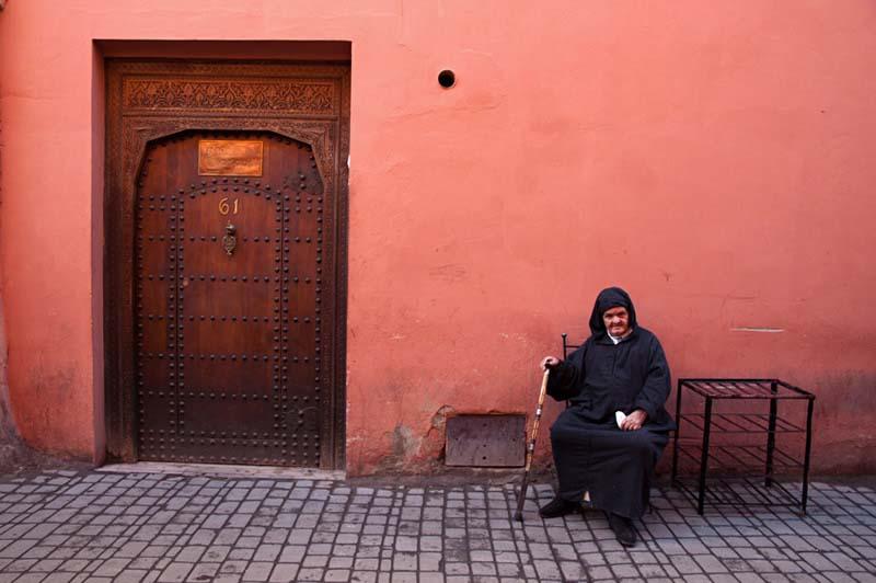 پیرزنی مراکشی با عصا نشسته کنار ساختمانی قرمز رنگ